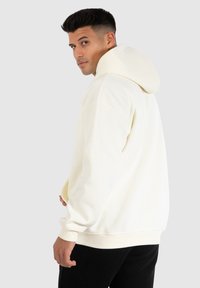 Gräddfärgad hoodies med stor huva, ribbade ärmslut och avslappnad passform, i kombination med svarta byxor. Mjuk tyg, minimalistisk design, inga synliga mönster.