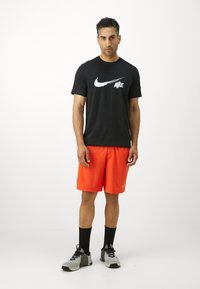T-shirt Nike noire avec logo blanc, short de sport orange, chaussettes noires et baskets grises avec motif texturé et accents noirs.