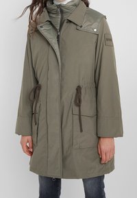 Kvinna som bär en olivgrön parkas med framfickor, justerbara midjekordlar och en synlig dragkedja samt tryckknappar.