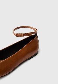 Chaussure plate en cuir marron avec une fine bride à la cheville et une boucle dorée, comportant un petit détail de croix dorée sur la bride sur fond gris clair.