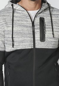 Felpa con zip grigia e nera con un design mélange, dotata di tasche con zip frontali e una piccola tasca sul petto con logo. Tessuto morbido e testurizzato.