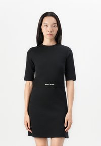 Rochie neagră, slim fit, cu mâneci scurte și decolteu înalt. Prezintă o bandă cu logo la talie și un material neted, texturat.