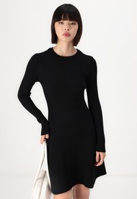Robe en tricot côtelé noire avec manches longues, col rond et ourlet évasé. Le matériau est doux avec une texture verticale. Accessoirisée avec un sac blanc.