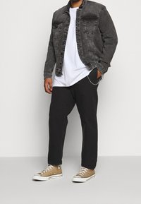 Uomo con giacca di jeans nera, t-shirt bianca, pantaloni neri con catena argentata e sneakers alte beige che sta in piedi contro uno sfondo bianco.