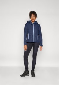 Marinewaterdichte jas met capuchon, contrasterende lichtblauwe rits, twee zijzakken, gecombineerd met zwarte leggings en wandelschoenen.