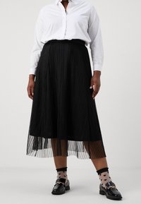 Svart plisserad midikjol med ett genomskinligt överdrag, med en texturerad design. Kombineras med en vit skjorta och svarta loafers med gulddetaljer.