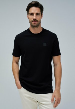 Camiseta de manga corta negra hecha de un tejido texturizado, con un pequeño parche de logo cuadrado en el pecho. Cuello redondo clásico.