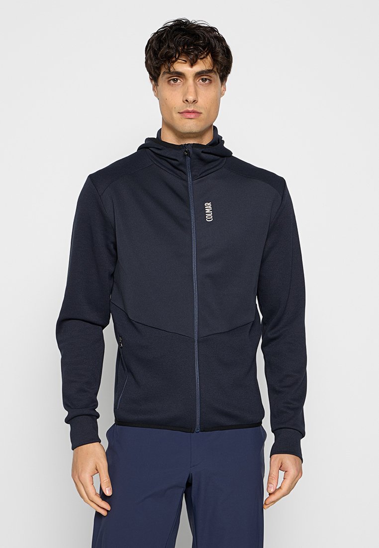 Giacca zip-up blu scuro realizzata in tessuto morbido. Presenta un cappuccio, due tasche laterali e un logo sulla zona sinistra del petto.