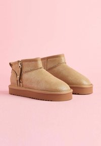 Botas de tobillo brillantes en tono bronce con punteras redondeadas, cierre de cremallera en el lateral y una suela de goma texturizada, colocadas sobre un fondo rosa.