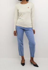 Cremfarbene und olivgrüne gestreifte Langarmbluse mit einem kleinen roten Herzdetail auf der Brust, kombiniert mit hellblauen geraden Jeans und schwarzen High Heels.
