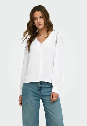 Jeune femme aux longs cheveux ondulés portant un chemisier blanc boutonné avec des détails en dentelle sur les manches et un jean bleu taille haute.
