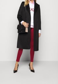 Donna che indossa un lungo cappotto nero, una felpa bianca con testo rosso, pantaloni rossi, tacchi neri e porta una borsa a tracolla nera.