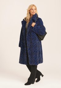 Long blue leopard-print coat made of soft fabric, featuring a wide collar, double-breasted buttons, and side pockets, paired with black pants.