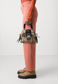 Bolso de mano marrón con cordones negros y un acento de hardware plateado. Superficie texturizada y colores contrastantes; sostenido por una persona que lleva pantalones rosas y zapatos marrones.