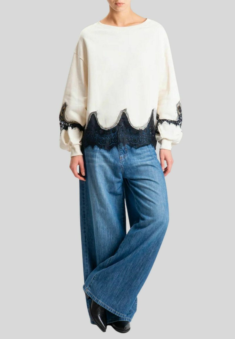 Felpa color crema con bordo in pizzo nero, caratterizzata da orli ondulati; abbinata a jeans in denim blu a gamba larga; finitura liscia e strutturata.
