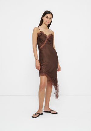 Femme portant une robe asymétrique marron en satin sans manches avec bordure en dentelle et des tongs noires, debout devant un fond blanc.
