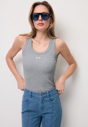 Vrouw met oversized zwarte zonnebril, grijze mouwloze top met ribstructuur en blauwe high-waist jeans, poseert met handen in de zij tegen een witte achtergrond.