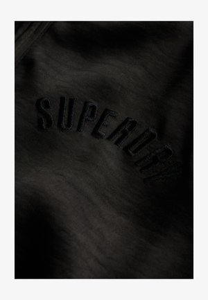 Sort stof med broderet "SUPERDRY" tekst i en matchende sort farve, der fremhæver en glat tekstur og subtil glans.