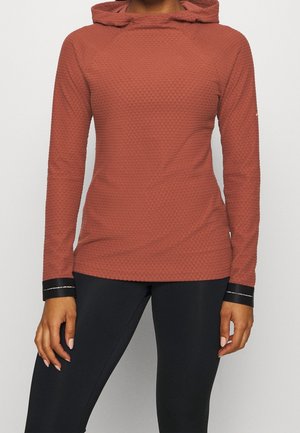 Mujer con sudadera con capucha de manga larga texturizada de color óxido y leggings negros, de pie con las manos relajadas a los lados.