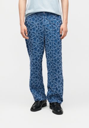 Blå bukser med store teksturerte løkkede blomster mønstre, brukt med svarte sko og en lyseblå skjorte, mot en ensfarget lys bakgrunn.