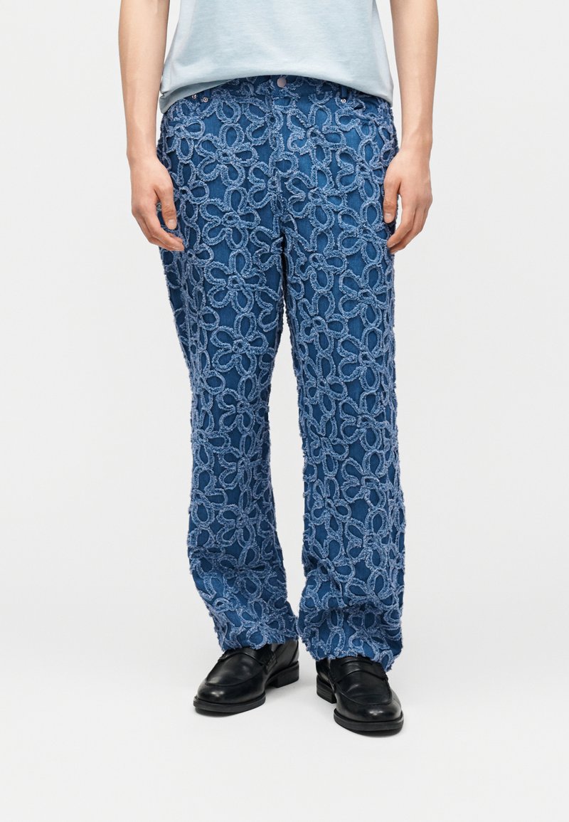 Μπλε παντελόνι με μεγάλα υφασμένα λουλουδένια σχέδια με θηλιές, συνδυασμένο με μαύρα παπούτσια και ανοιχτό μπλε πουκάμισο, πάνω σε απλό φωτεινό φόντο.