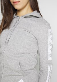 Felpa grigia con zip, cappuccio e tasche frontali. Le maniche presentano il logo Adidas bianco e un motivo a tre strisce. Realizzata in tessuto morbido.