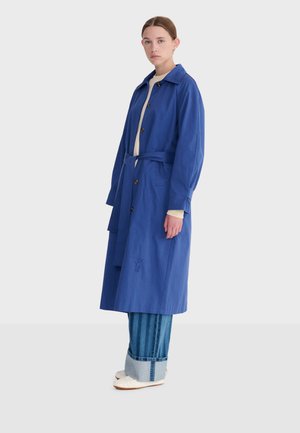 Kvinde iført en lang blå kåbe med bælte over opklebede jeans og hvide sko, stående mod en ensartet lys baggrund.