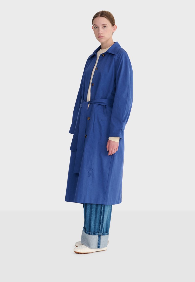 Femme portant un long manteau bleu ceinturé sur un jean retroussé et des chaussures blanches, debout devant un fond clair uni.