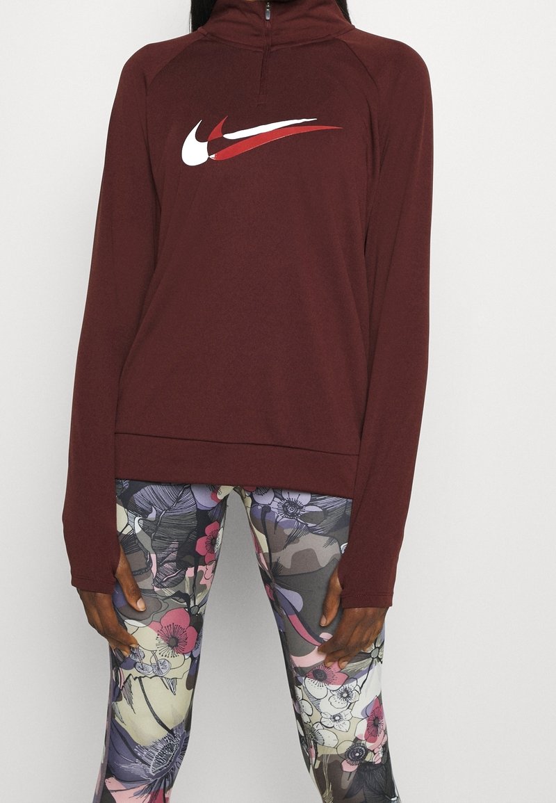 Maronfarbener Sweatshirt mit Reißverschluss, der auf der Vorderseite ein weißes und rotes Nike-Logo aufweist, kombiniert mit gemusterten Leggings in Pastellfarben mit floralen Motiven.