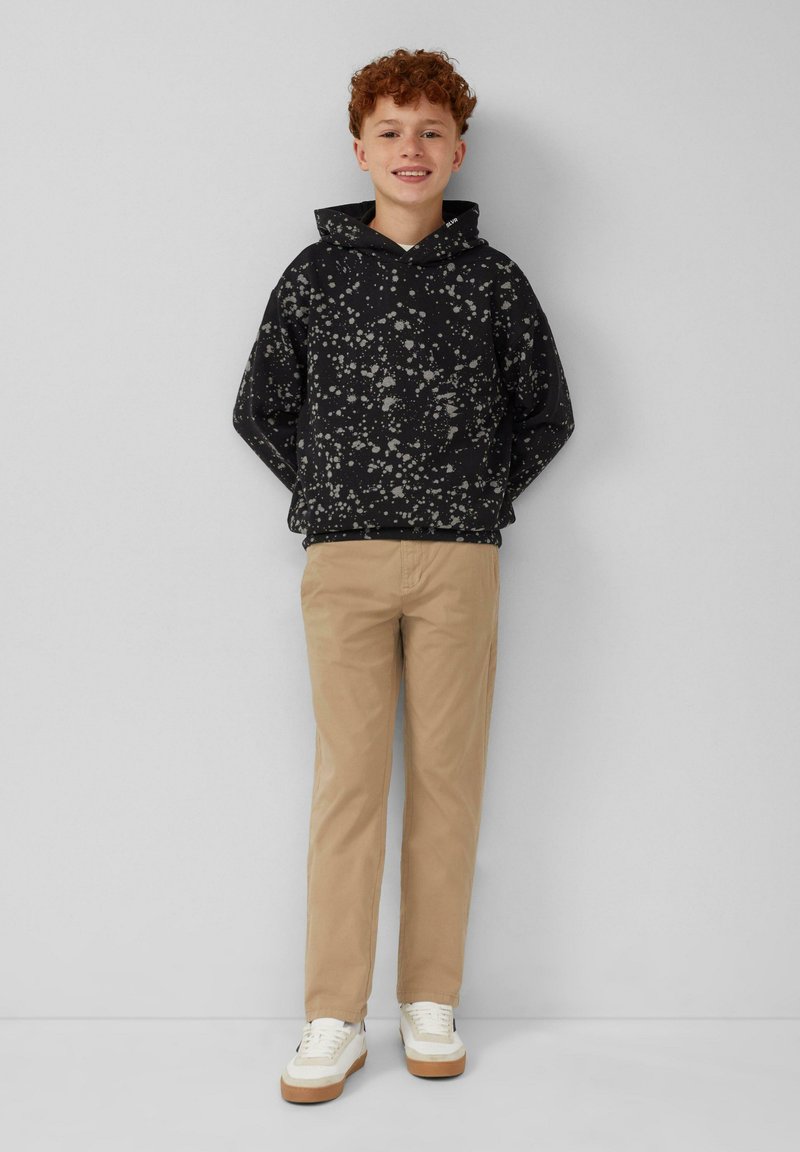 Glunderende jongen met krullend rood haar, gekleed in een zwart gespetterde hoodie, beige broek en witte sneakers met gumzolen, staand tegen een grijze muur.