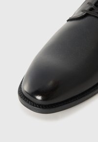 Black leather shoe with a polished finish, pointed toe, and stitched detailing along the sole. Minimalist design with laces.