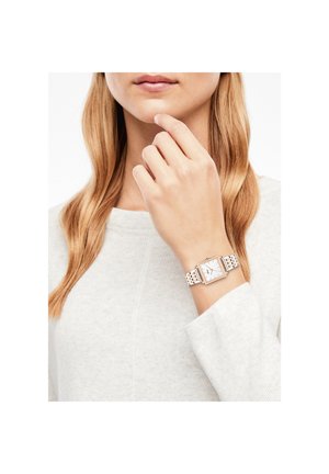 Femme aux cheveux longs et blonds portant un pull blanc et une montre-bracelet carrée argent et or au poignet gauche.