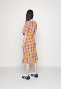 Vestido xadrez com cores vibrantes amarelo, rosa e roxo, mangas curtas, um recorte nas costas e uma bainha larga. Estilizado com ténis robustos.