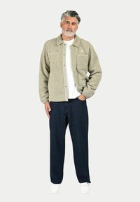 Giacca in pile beige con chiusura a bottoni, due tasche frontali, indossata sopra una camicia bianca e pantaloni in denim scuro, abbinata a sneakers bianche.