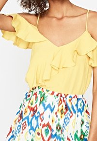 Top giallo senza maniche con maniche arricciate e scollatura a V, abbinato a una gonna a fantasia colorata con disegni geometrici blu, rossi e verdi.