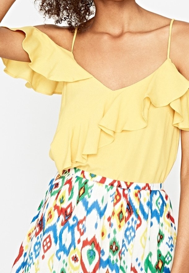 Top giallo senza maniche con maniche arricciate e scollatura a V, abbinato a una gonna a fantasia colorata con disegni geometrici blu, rossi e verdi.
