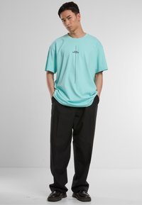 Tricou verde mentă cu mânecă scurtă și guler rotund, cu text negru și un design cu linii verticale. Asortat cu pantaloni negri lejeri și pantofi închisi la culoare.