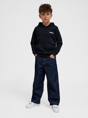 Drenge med krøllet hår iført en sort hættetrøje med "Hummel" logo, brede mørke jeans og hvide sneakers, stående med hænderne i lommerne.