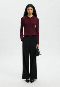 Nais, kellel on lokkis juuksed, kannab burgundia värvi pikkade varrukatega nööbitoppimist, musti laienevaid pükse, musti terava ninaga kingi ning hoiab käes väikest musta käekotti.