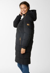 Woman smiling and wearing a long black padded coat with hood, standing in profile with hand near neck against white background.