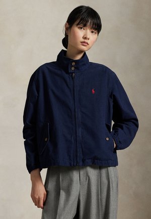 Une femme porte une veste bleu marine zippée à col montant, ornée d’un logo rouge sur la poitrine, avec un pantalon gris, sur un fond uni.