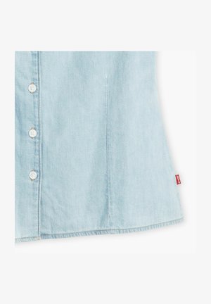 Lyseblå denimskjorte med knaplukning fortil, hvide knapper og buet bund. Har en rød mærkat ved sømmen.