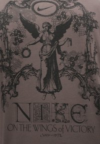 Γκρίζα μπλούζα με κοντό μανίκι, με μαύρο εκτυπωμένο σχέδιο που απεικονίζει έναν άγγελο που κρατάει ένα στεφάνι, περιτρυγιρισμένο από περίπλοκα μοτίβα και το λογότυπο της Nike.