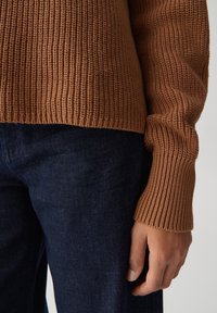 Person, der einen braunen gerippten Strickpullover trägt, der in dunkle blaue Jeans gesteckt ist, mit einer entspannten Hand an der Seite.