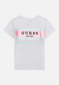 Bela kratka majica s kratkimi rokavi, ki vsebuje obarvane vodoravne črte in črn napis "GUESS EST.1981" z dekorativnimi motivi v pastelnih barvah.