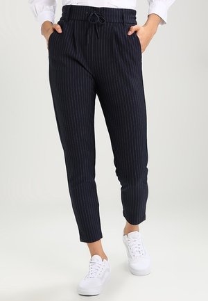 Osoba ubrana w czarne spodnie o długości do kostek w cienkie prążki ze ściągaczem w talii, białe trampki i białą, długorękawową koszulę włożoną do środka, z rękami w kieszeniach.