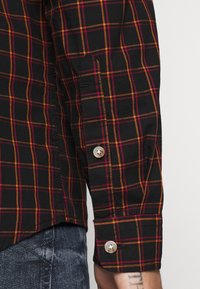 Schwarzes karierter Shirt mit orangefarbenen und burgunderfarbenen Linien, mit Knopfmanschetten und einem strukturierten Material. Sichtbare Steppnähte am Ärmel.