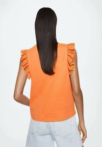 Haut orange sans manches avec épaules à volants, en tissu doux. Le dos est uni avec un col rond, associé à un short en jean clair.