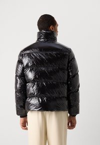 Giacca a vento nera con un esterno lucido e trapuntato, colletto alto e sezioni cucite orizzontalmente, indossata sopra pantaloni chiari.