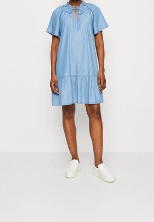 Kobieta w jasnoniebieskiej sukience z krótkim rękawem, z marszczonym dołem i wiązaniem przy dekolcie, w białych wsuwanych trampkach, stojąca na jednolitym tle.